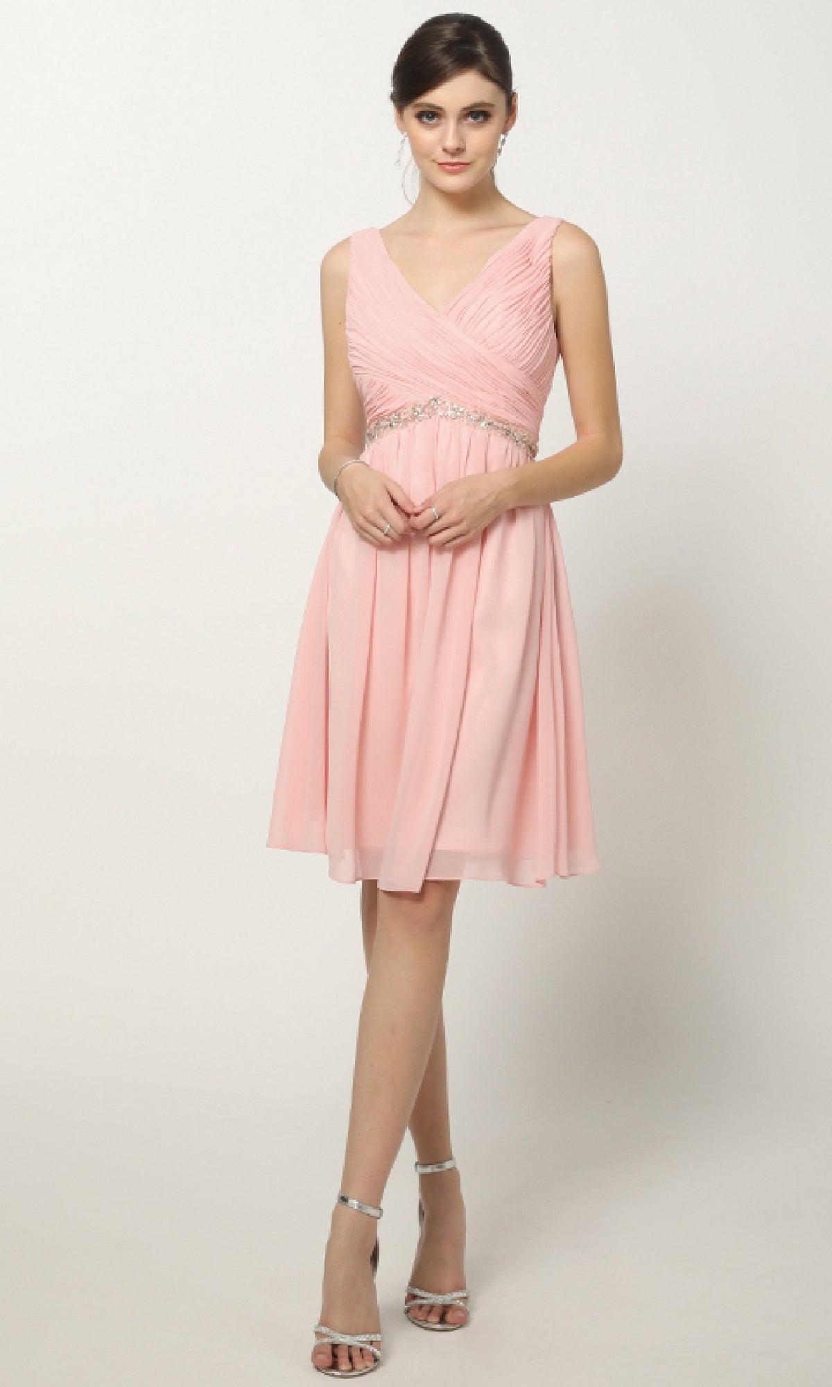 A woman stands against a plain background wearing the Juliet 786, a sleeveless, knee-length pink semi-formal dress with a V-neckline, paired with silver high-heeled sandals.